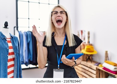 Young blonde woman working as manager at retail boutique crazy and mad shouting and yelling with aggressive expression and arms raised. frustration concept. 
