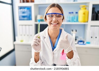 Young Blonde Woman Working At Scientist Laboratory Holding Sample Smiling Happy And Positive, Thumb Up Doing Excellent And Approval Sign 