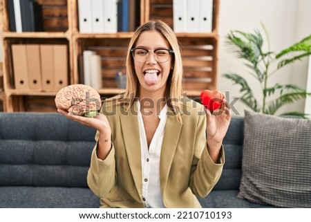 Young blonde woman working at therapy office holding brain and health sticking tongue out happy with funny expression. 