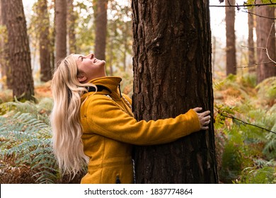 Young Blonde Woman In Yellow Coat Hugging A Tree In The Forest And Looking Up To The Tree Top