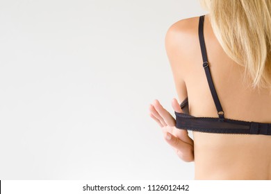 Young Blonde Woman's Hand Putting On Or Taking Off A Black Bra. Choosing Right Size.  Daily Underwear Concept. View On The Back. Empty Place For Text On The Light Gray Background.