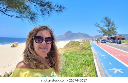 The young blonde in a yellow blouse and sunglasses poses smiling on a sunny morning with the sea and Pedra do Elefante (Elephant Rock) in the background. Itaipuaçu, Maricá, Rio de Janeiro.