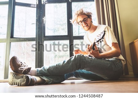 Young blondhair handsom man plays on guitar sitting on floor near the whole wall window at home. Funny freetime spending concept image.