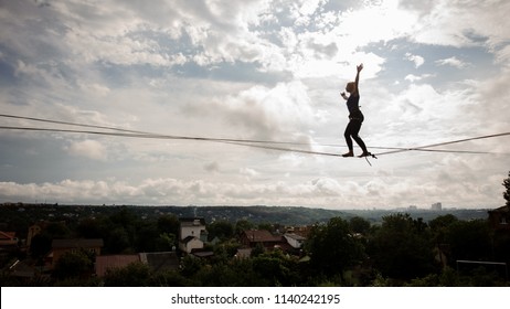 Young blondie woman walking on the slackline rope on the bending knees on the background of houses among trees and clear sky