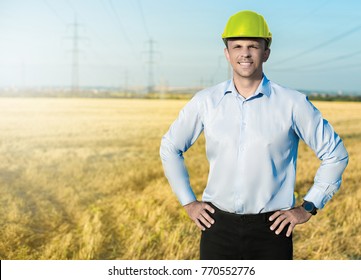 Young Blue Collar Worker Or Engineer Wearing In Yellow Helmet Stands In The Field With Wide Smile. Half-length Portrait.