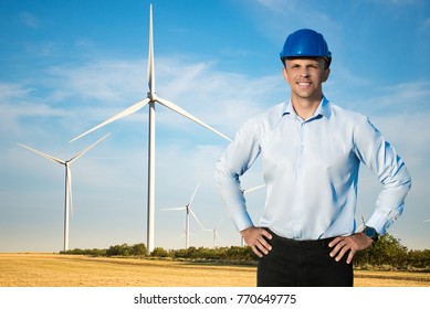 Young Blue Collar Worker Or Engineer Wearing In Yellow Helmet Stands In The Field With Wide Smile Against Wind Turbines Farm. Half-length Portrait.