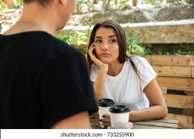 Young bored girl sitting and drinking coffee with her boyfriend at a cafe outdoors