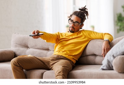Young Bored Tired African American Man Pressing Button On Remote Control While Watching Television At Home After Hard Working Day, Black Guy Sitting On Sofa With Popcorn Bowl And Changing Tv Channel