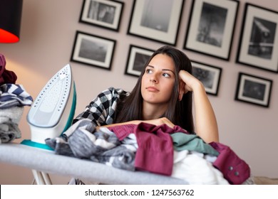 Young Bored And Tired Woman Leaning On The Ironing Board, Surrounded With Bunch Of Wrinkled Clothes Ready For Ironing