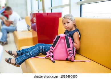 Young Boring Girl Sitting In Airport Lounge On Sofa With Her Small Pink Bagpack, Waiting For A Flight