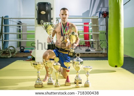 Young boxer champion with many medals around his neck posing below his trophy cups in sports hall.
