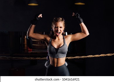 Young boxer girl with boxing gloves screams emotionally rising arms in victory sign showing fit and healthy body in female fighter and successful woman concept. Loft gym ring with ropes.