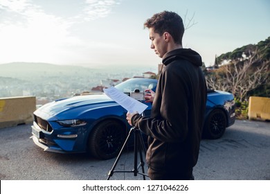 Young Boy With Black Sweatshirt Reading Script And Preparing Camera To Record Video On Viewpoint With Blue Luxury Car And Sporty