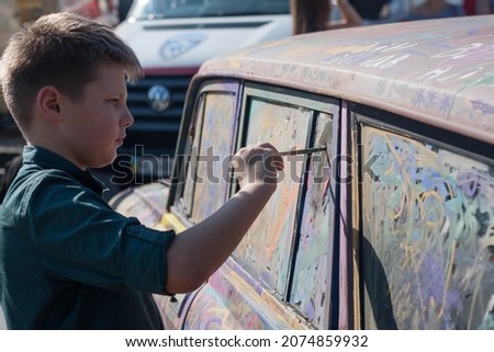A young boy with a brush for drawing in his hands paints a car in bright paints. Street art.