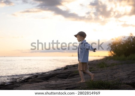 Young boy in a cap and denim jacket strolls barefoot on rocky shoreline during sunset, with water and clouds visible in the background