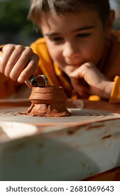 Young boy carefully shaping red clay on a pottery wheel using a loop tool, enjoying a creative outdoor activity