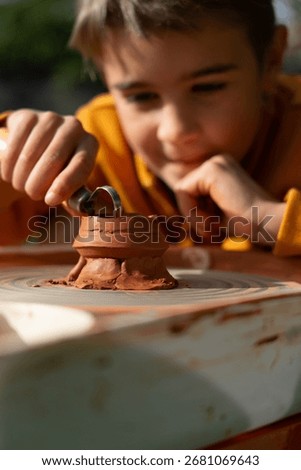 Young boy carefully shaping red clay on a pottery wheel using a loop tool, enjoying a creative outdoor activity
