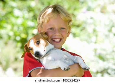 Young boy carrying his dog in his arms and smiling at the camera on a bright, sunny day in the park