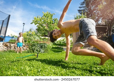 A young boy cartwheeling joyfully on a sunlit lawn with water sprinkles around as another child watches from behind - Powered by Shutterstock