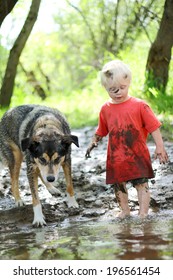 A Young Boy Child And His German Shepherd Mix Dog Are Covered In Mud And Playing Outside On The Beach Of A River In The Woods.