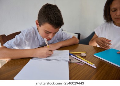 A young boy concentrates on his homework at a table, with school supplies scattered around, while a parent provides support and guidance nearby. - Powered by Shutterstock