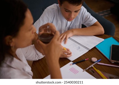 A young boy concentrates on his math homework, jotting down notes in a spiral notebook. An adult, possibly a parent or tutor, sits nearby, providing support and guidance. - Powered by Shutterstock