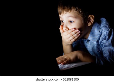 Young Boy Covered Her Mouth In Fright, Sitting At A Computer On A Dark Night. The Child Is Shocked By What He Saw On The Laptop. Watching A Scary Movie.