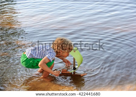 Young boy crouches down in shallow calm lake water and blows on the sail of his homemade sailing boat. It is summertime.