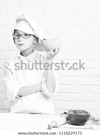 young boy cute cook chef in white uniform and hat on stained face flour with glasses standing near table with rolling pin and holding turquoise bowl with chocolate cookies on brick wall background.