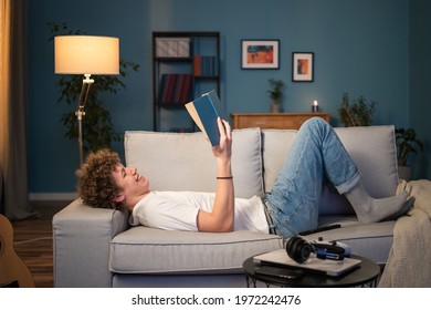 A young boy with dark curly hair lies on a couch in the living room in homespun clothes leaning against the back of the sofa. The teenager is relaxing reading a book, studying, laughing