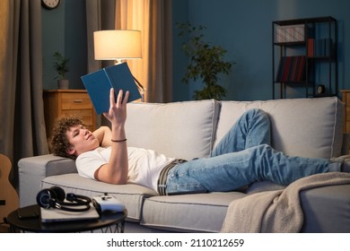 A young boy with dark curly hair lies on a couch in the living room in homespun clothes leaning against the back of the sofa. The teenager is relaxing by reading a book, studying - Powered by Shutterstock