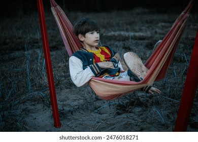 A young boy with dark hair relaxes in a striped hammock wearing a colorful jacket and sneakers. He is outside on the sandy ground with a contemplative expression on his face.  - Powered by Shutterstock