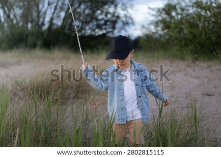 Young boy in a denim jacket and cap stands among tall grass at the riverbank, holding a stick and engaging with the natural surroundings