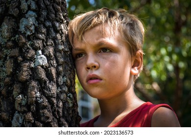 A young boy with a dirty sweaty face leaning his head against a wild persimmon tree- A little boy with a serious expression on his face, thinking about something while leaning his head against a tree