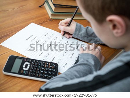 Young boy doing maths homework test times tables multiplication exam paper sat at table homeschooling education with calculator and books holding pencil learning mathematics  getting answers right