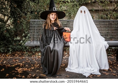 Young boy dressed in a bed sheet to be a ghost, and a young girl in witch costume holding a pumpkin pail for halloween.