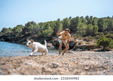 A young boy dressed in clothes, throwing a stick to his dog on the seashore of a Mediterranean beach.