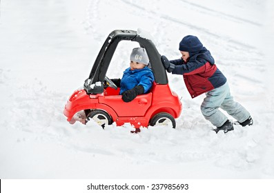A young boy dressed for cold weather sits in a red toy car stuck in the snow during the winter season.  His older brother helps by giving the car a push from behind. 