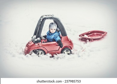 A Young Boy Dressed For Cold Weather Sits In A Red Toy Car Stuck In The Snow Pulling Behind A Red Sled During The Winter Season.  Filtered For A Retro, Vintage Look. 