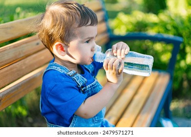 Young Boy Drinking Water on Bench. A young boy in blue overalls sits on a wooden bench and drinks from a clear plastic water bottle. - Powered by Shutterstock