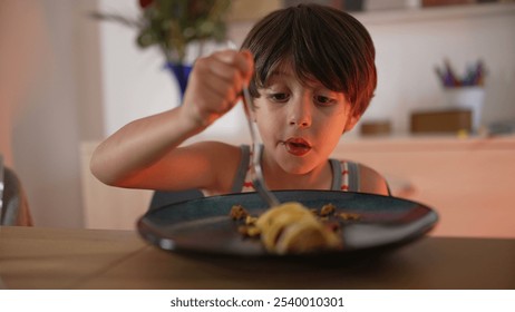 Young boy eating at the dining table, using a fork to take food from his plate, demonstrating focus and enjoyment during meal time in a home environment - Powered by Shutterstock