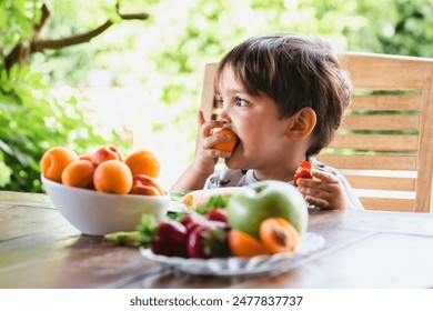 Young boy enjoying fresh fruit at a table in the garden, surrounded by vibrant greenery and various fruits. - Powered by Shutterstock