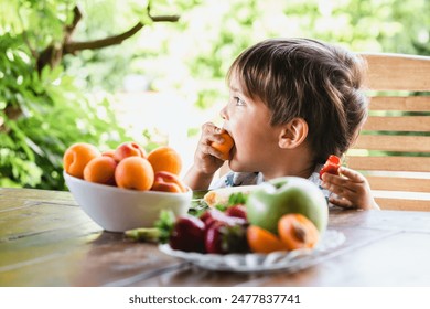 Young boy enjoying fresh fruit at a table in the garden, surrounded by vibrant greenery and various fruits. - Powered by Shutterstock