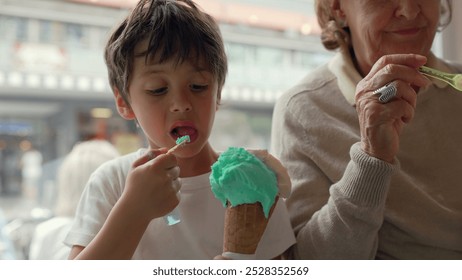 Young boy enjoying a vibrant ice cream cone, taking a big bite while sitting next to his grandmother in a lively indoor setting. sweet moment of intergenerational bonding over a treat - Powered by Shutterstock