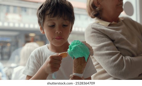 Young boy enjoying a vibrant ice cream cone, taking a big bite while sitting next to his grandmother in a lively indoor setting. sweet moment of intergenerational bonding over a treat - Powered by Shutterstock