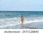 A young boy enjoying the waves at the beach, walking along the shoreline with the vast blue ocean stretching out to the horizon under a clear sky.