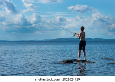 The young boy fishing by the spinning from the stone shore of the big lake. The kid trying catch a fish by himself. The topless boy fishing in the fresh water. A teenager throws a spinning rod  - Powered by Shutterstock