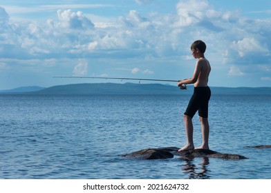 The young boy fishing by the spinning from the stone shore of the big lake. The kid trying catch a fish by himself. The topless boy fishing in the fresh water. A teenager throws a spinning rod  - Powered by Shutterstock