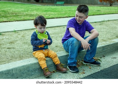 Young boy with flat top haircut in a purple t-shirt and jeans sitting on a street curb with his toddler brother - Shutterstock ID 2095548442