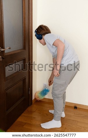 A young boy is focused on cleaning a corner of the room using a vibrant duster. He wears headphones and stands on a wooden floor near a closed door.